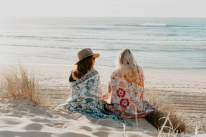 Two girls watching sunrise on beach with pink and green floral florence throw wrapped around them