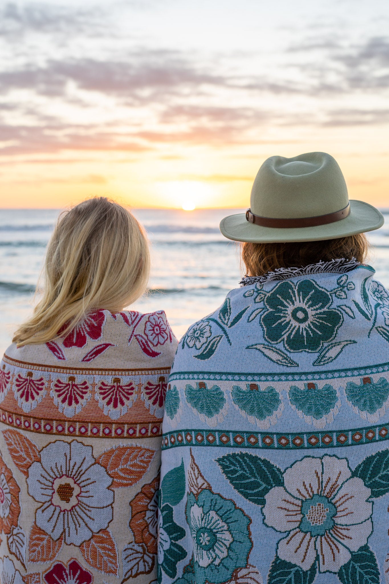 Girl & boy watching sunrise on beach with pink and green floral florence throw wrapped around her